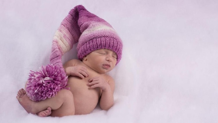 Full length of 2 weeks old newborn baby girl sleeping on stomach wearing a purple knitted hat.