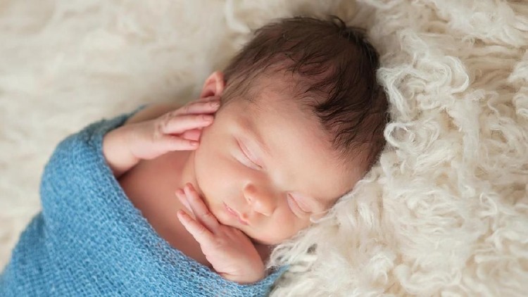 A 12 day old newborn baby boy sleeping on a beige flokati rug and swaddled in a gauzy light blue wrap.