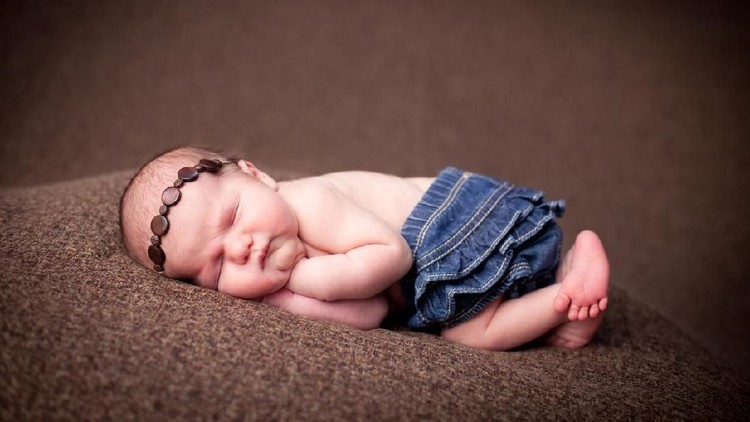 Color image of a newborn baby girl wearing a beaded headband while sleeping peacefully on brown, textured fabric.  With copy space.