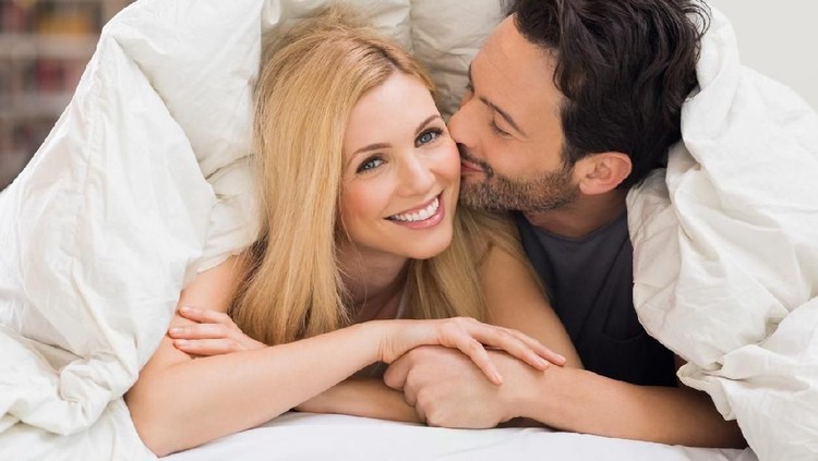 Handsome man kissing woman on cheek under a duvet in the bed. Happy young couple lying down comfortably in their bed at home. Young couple having fun lying on bed at home.