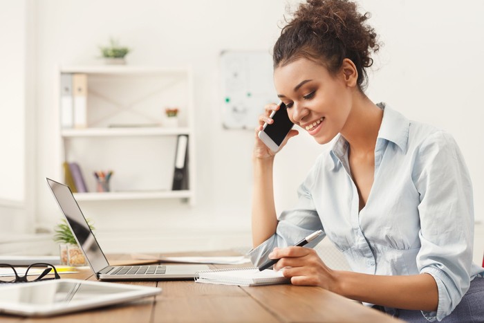 Smiling african-american business woman at work talking on phone and taking notes, sitting at her working place in office, copy space