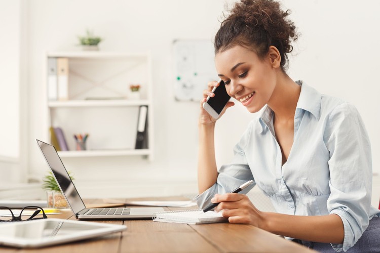 Smiling african-american business woman at work talking on phone and taking notes, sitting at her working place in office, copy space