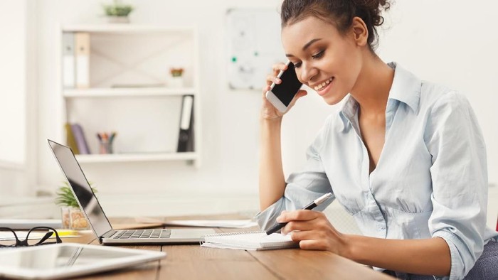 Smiling african-american business woman at work talking on phone and taking notes, sitting at her working place in office, copy space