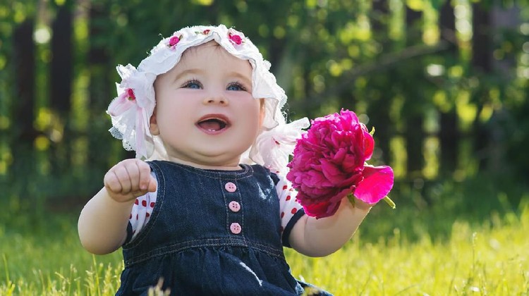 cute little baby girl sitting on the grass with flower in her hand on the nature, outdoor. child with big blue eyes laughing in a summer hat