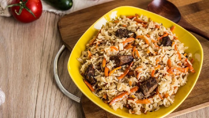 Photo of dish of uzbek pilaf made of rice and carrots, meat and onions. Close-up photo on a wooden background with a spoon and tomato