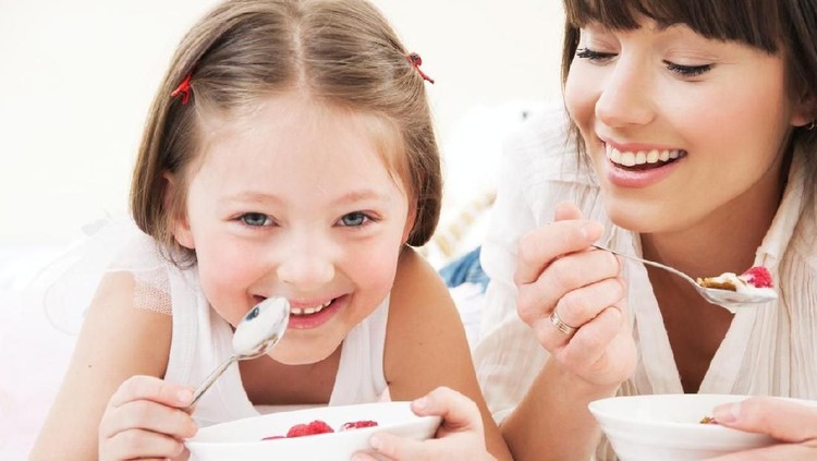 Happy young family, mother with two children, adorable toddler girl and funny messy baby boy having healthy breakfast eating fruit and dairy, sitting in a white sunny kitchen with window