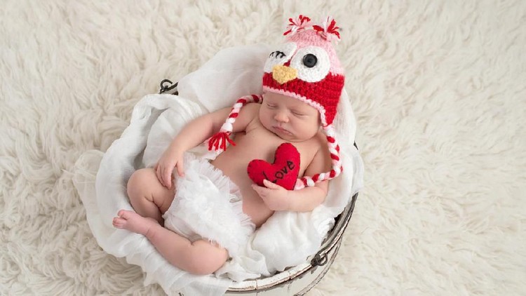 A nine day old, newborn baby girl wearing a pink and red owl hat and holding a heart shaped pillow with the word, 