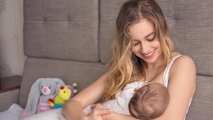 Breastfeeding baby. Pretty mother holding her newborn child. Mom smile and nursing infant. Beautiful woman and new born love at home. Blond mother breast feeding baby.