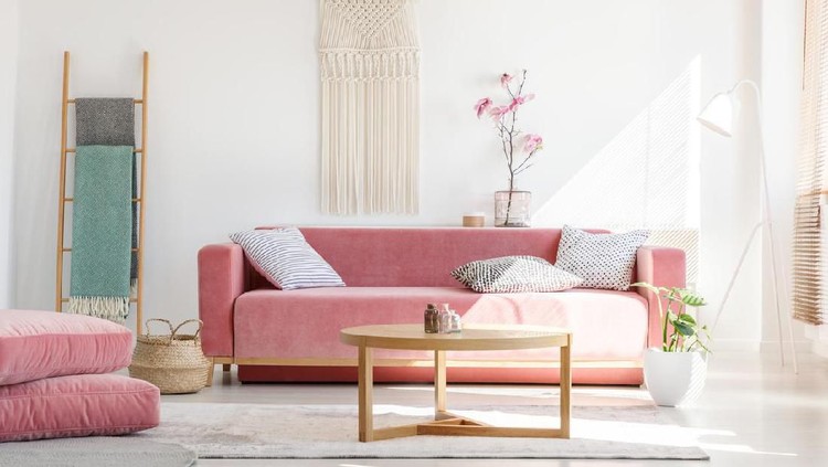 Cozy cushions and stylish textiles in a sunny, feminine living room interior with a pink, velvet sofa and a macrame on a white wall