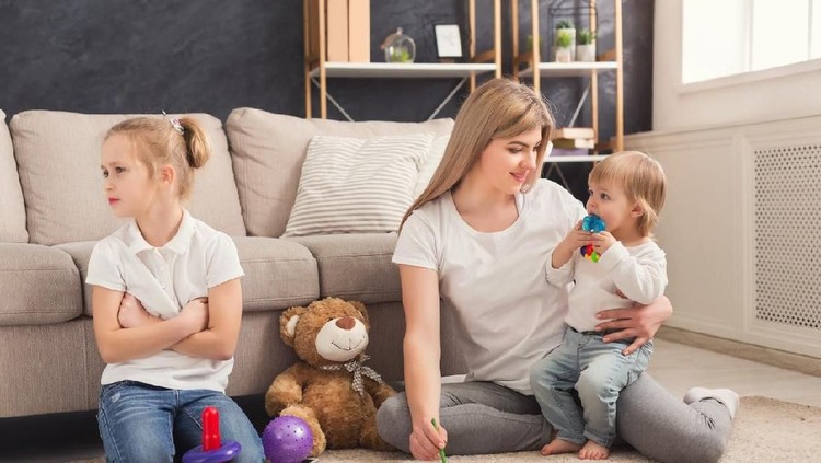 Happy mother playing with her little child while elder daughter feeling offended. Relationship, family issues and motherhood concept
