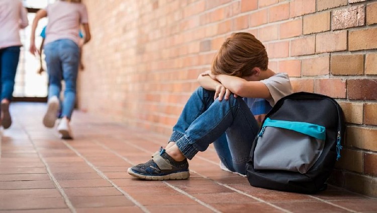 Little boy sitting alone on floor after suffering an act of bullying while children run in the background. Sad young schoolboy sitting on corridor with hands on knees and head between his legs.