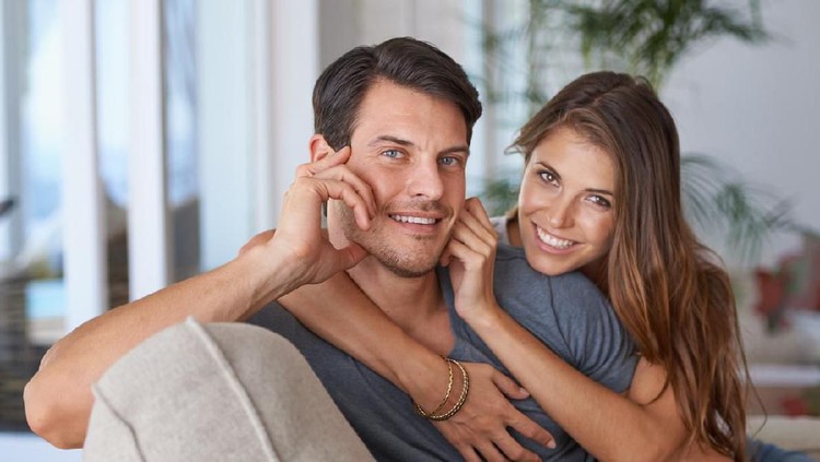 Young tender husband hugging his wife, loving man embracing woman gently and tightly sitting on couch at home, showing love and support, happy soulmates or close friends, peace, harmony, understanding