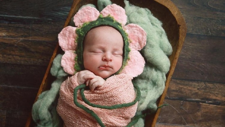 A newborn baby girl, fast asleep, is wearing an adorable flower bonnet.