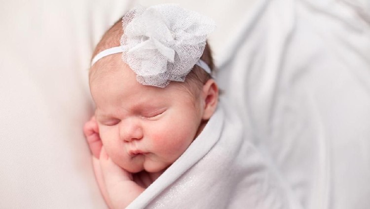 Color photo of a newborn baby girl wearing a headband and  sleeping peacefully, covered with a white blanket.