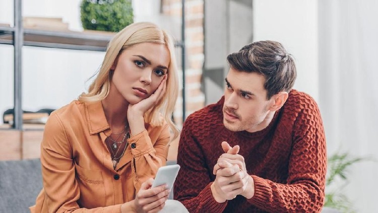 husband looking at unhappy young woman holding smartphone and looking at camera at home, relationship problem concept