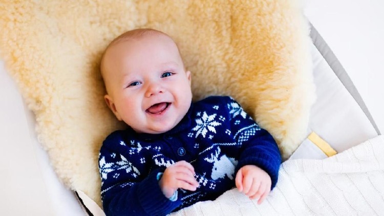 Funny little baby relaxing in a bouncer with sheepskin foot muff wearing warm knitted reindeer Christmas sweater at home on cold winter day. New born boy in stroller. Newborn child in rocking chair.