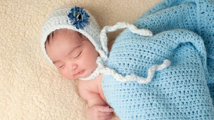 A 7 day old, sleeping Hispanic baby girl wearing a light blue, crocheted bonnet with flower decoration. She is covered with a blue crocheted blanket and sleeping on a beige blanket.