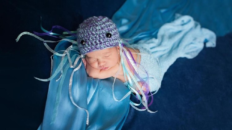 Six day old newborn baby girl wearing a crocheted jellyfish hat and sleeping on blue fabric.