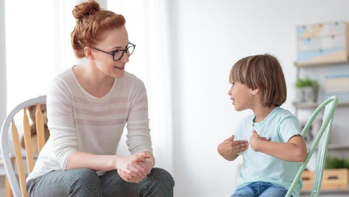 Smiling little boy sitting on chair talking with cheerful child psychotherapist during therapy session at office