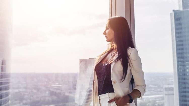 Portrait of elegant business lady wearing white formal suit standing near window looking at cityscape.