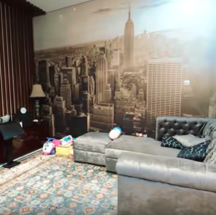 <p>Terasa kontras dengan ruang keluarga yang berhadapan langsung. Di sini ditata sofa berwarna abu-abu yang berhadapan dengan sebuah tv besar. Menariknya, dipasang wallpaper sky view. Menurut Ussy mereka ingin menciptakan pemandangan seperti tingga di sebuah apartement. (Foto: YouTube Atta Halilintar)</p>