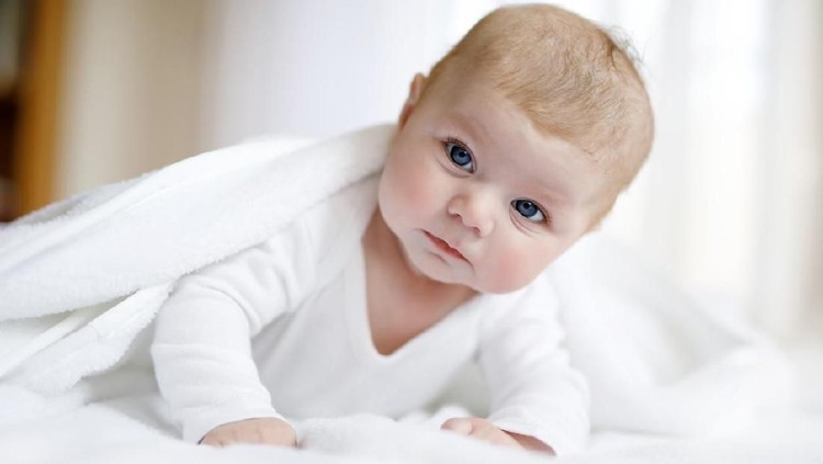 Baby girl with blue eyes wearing white towel or winter overal in white sunny bedroom. Newborn child relaxing in bed. Nursery for children. Textile and bedding for kids. New born kid with toy bear.