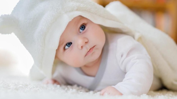 Baby girl with blue eyes wearing white towel or winter overal in white sunny bedroom. Newborn child relaxing in bed. Nursery for children. Textile and bedding for kids. New born kid with toy bear.
