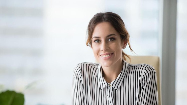 A smiling young businesswoman sits opposite an unrecognizable candidate during an interview.  She reaches across her desk to shake hands with him.