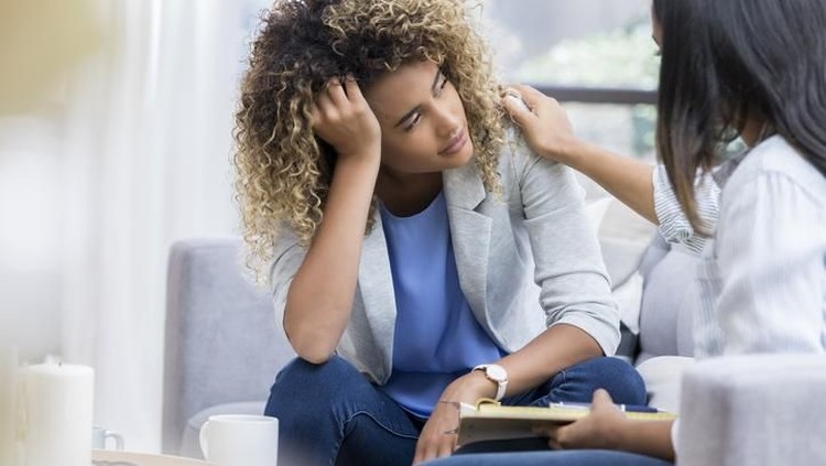A young woman sits on a couch with her unrecognizable therapist.  She puts her head in her hand as she looks out the window with a sad expression.  Her therapist puts a hand on her shoulder.