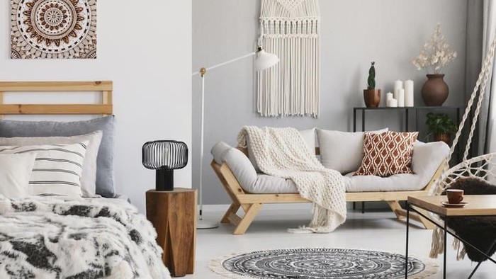 Small open space flat interior with beige sofa with cushion, macrame on the wall, rack with candles and plants and bed with pillows