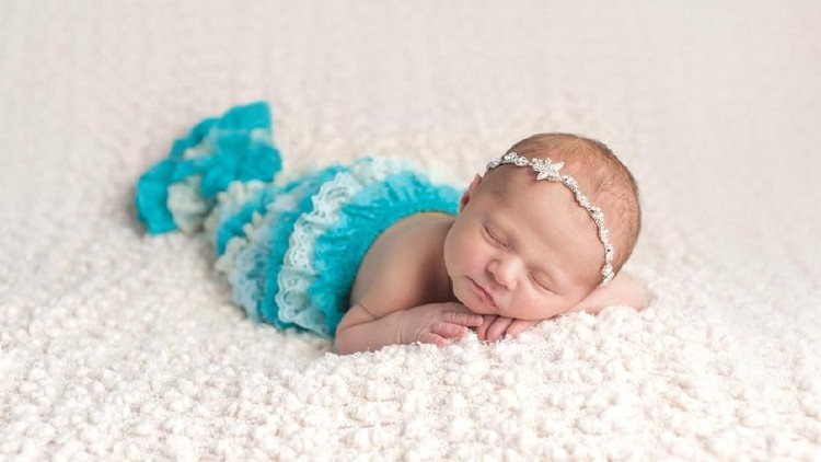 A sleeping, ten day old newborn baby girl wearing a lace aqua blue mermaid tail. She is lying on her stomach on a cream colored, bouncle blanket.