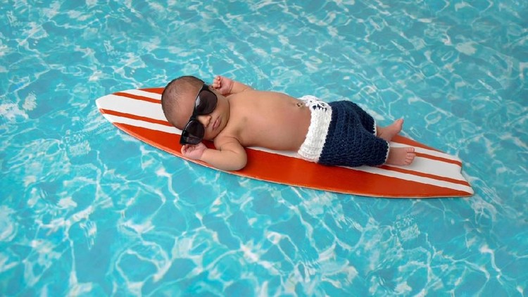 NB Nine day old newborn baby boy sleeping on a tiny, orange and white surfboard. He is wearing navy blue, crocheted board shorts and sunglasses.