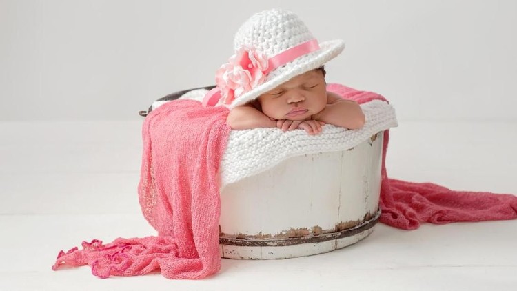 NB A portrait of a three week old, sleeping, newborn, baby girl wearing a fancy, brimmed Easter hat with pink flowers. She is sleeping in a vintage, white, wooden bucket.
