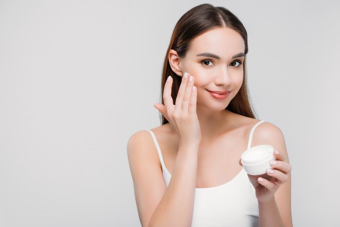 beautiful girl applying cosmetic cream on face, isolated on grey