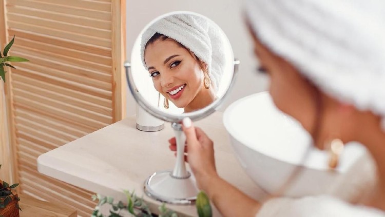 Attractive smiling woman with a white towel on her head dressed in bathrobe looks at herself in the mirror in stylish bathroom after morning shower. Beauty concept.