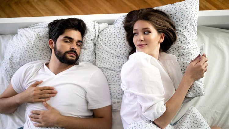 Beautiful young wife texting with lover on smartphone while husband is sleeping