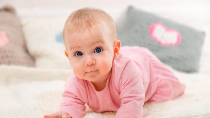 Charming beautiful lovely small female baby with plump cheeks creeps on bed, looks innocently on something, being naughty. Cute baby infant plays alone. Newborn kid crawls. Enthusiastic child