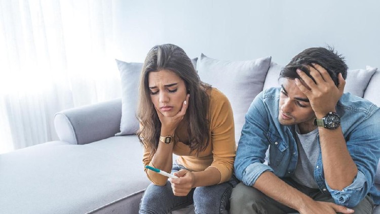 Sad young married couple embracing standing in living room opposite window at home. Sorrowful wife and husband feels unhappy, thinking about problems in relations, miscarriage, misbirth or infertility