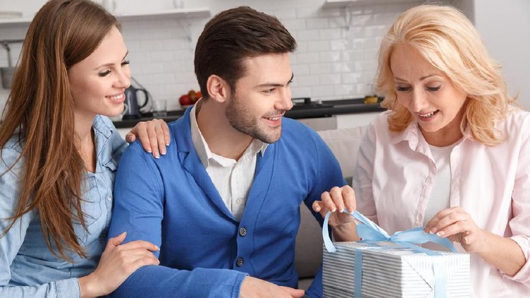 Young family man and woman with mother-in-law at home weekend sitting on sofa mother holding ribbons surprised ready to open box happy