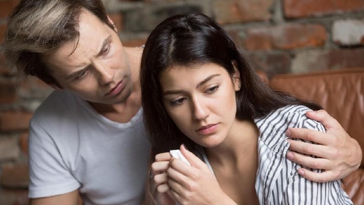 Suami dan istri Sad young married couple embracing standing in living room opposite window at home. Sorrowful wife and husband feels unhappy, thinking about problems in relations, miscarriage, misbirth or infertility