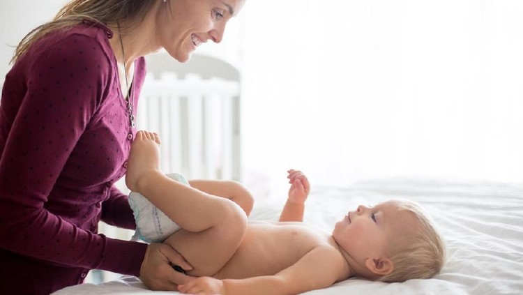 Young mom, changing baby diaper after bath in sunny bedroom