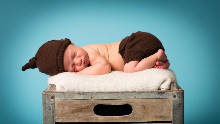 Color image of curled up, sleeping baby, wearing brown knit hat and diaper cover, lying on tummy, on an old, wood crate.