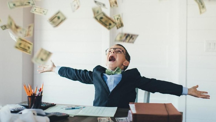 A young entrepreneur boy businessman is dressed in business attire and is working hard on his business while earnings in US currency are raining from the sky. He loves earning money from his new business and saving his money in the bank.