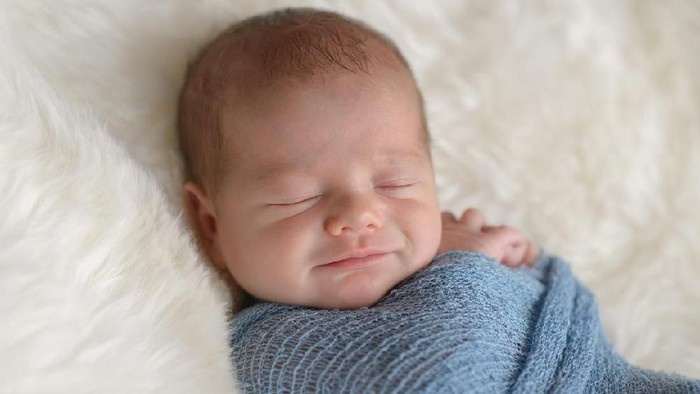 Sleeping, nine day old, newborn baby boy with a smile on his face. He is swaddled in a light blue wrap. Shot in the studio on a white, sheepskin rug.