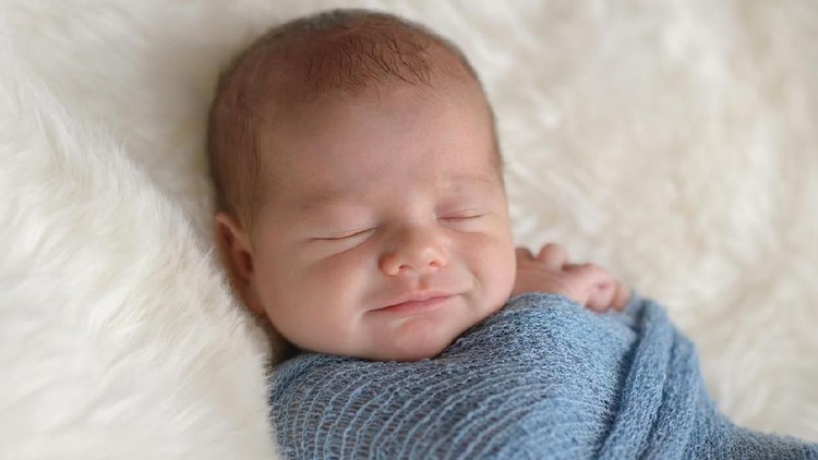 Sleeping, nine day old, newborn baby boy with a smile on his face. He is swaddled in a light blue wrap. Shot in the studio on a white, sheepskin rug.