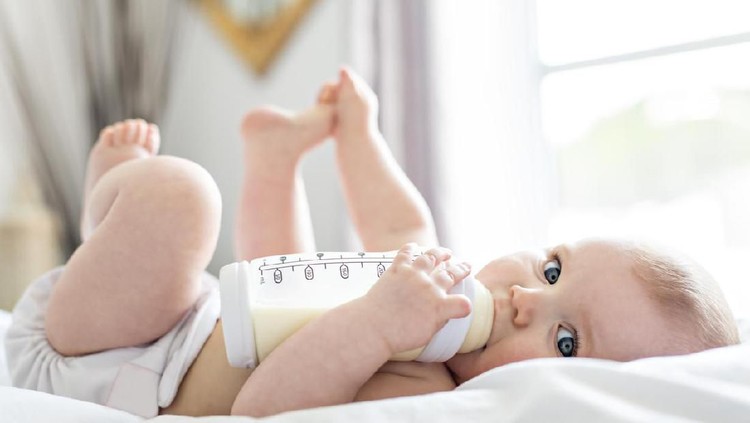 A Pretty baby girl drinks water from bottle lying on bed. Child weared diaper in nursery room.
