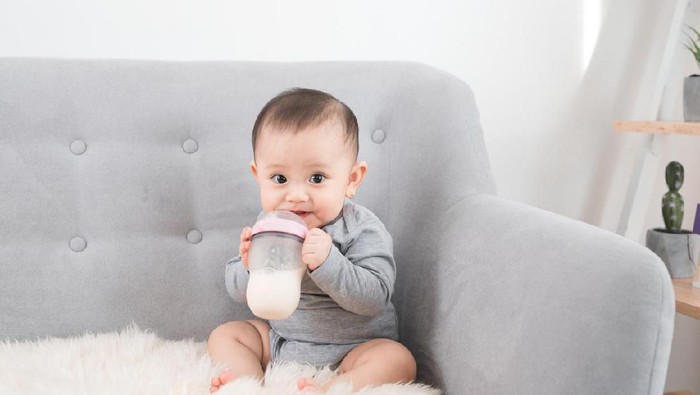 A Pretty baby girl drinks water from bottle lying on bed. Child weared diaper in nursery room.