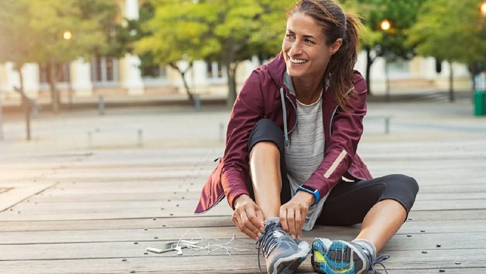 Mature fitness woman tie shoelaces on road. Cheerful runner sitting on floor on city streets with mobile and earphones wearing sport shoes. Active latin woman tying shoe lace before running.