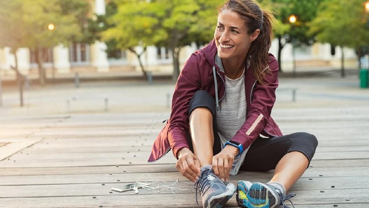 Mature fitness woman tie shoelaces on road. Cheerful runner sitting on floor on city streets with mobile and earphones wearing sport shoes. Active latin woman tying shoe lace before running.