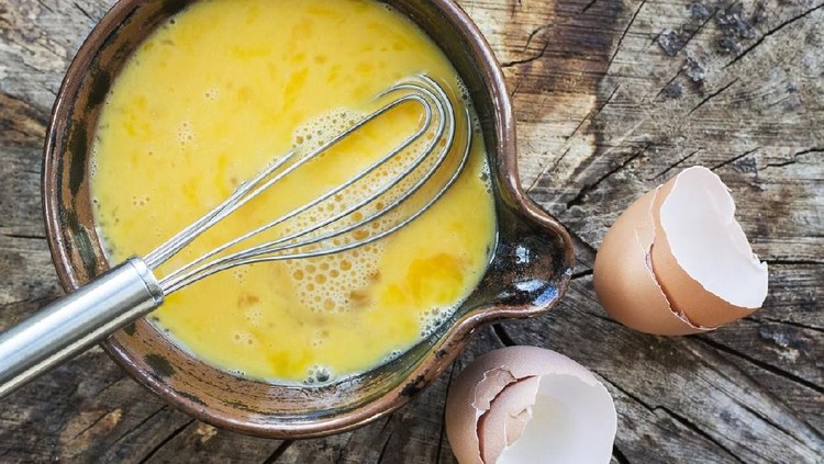 telur dadar Beaten eggs and whisk in a ceramic bowl. In this photograph the bowl of eggs is placed on a rustic wooden surface, the whisk is placed in the bowl of eggs and is projecting out of the photograph. The egg shells from the beaten eggs are placed on the wooden surface. This photograph is taken with natural light. The image is intended for chefs, cooks, restaurants and egg sellers, as well as other.